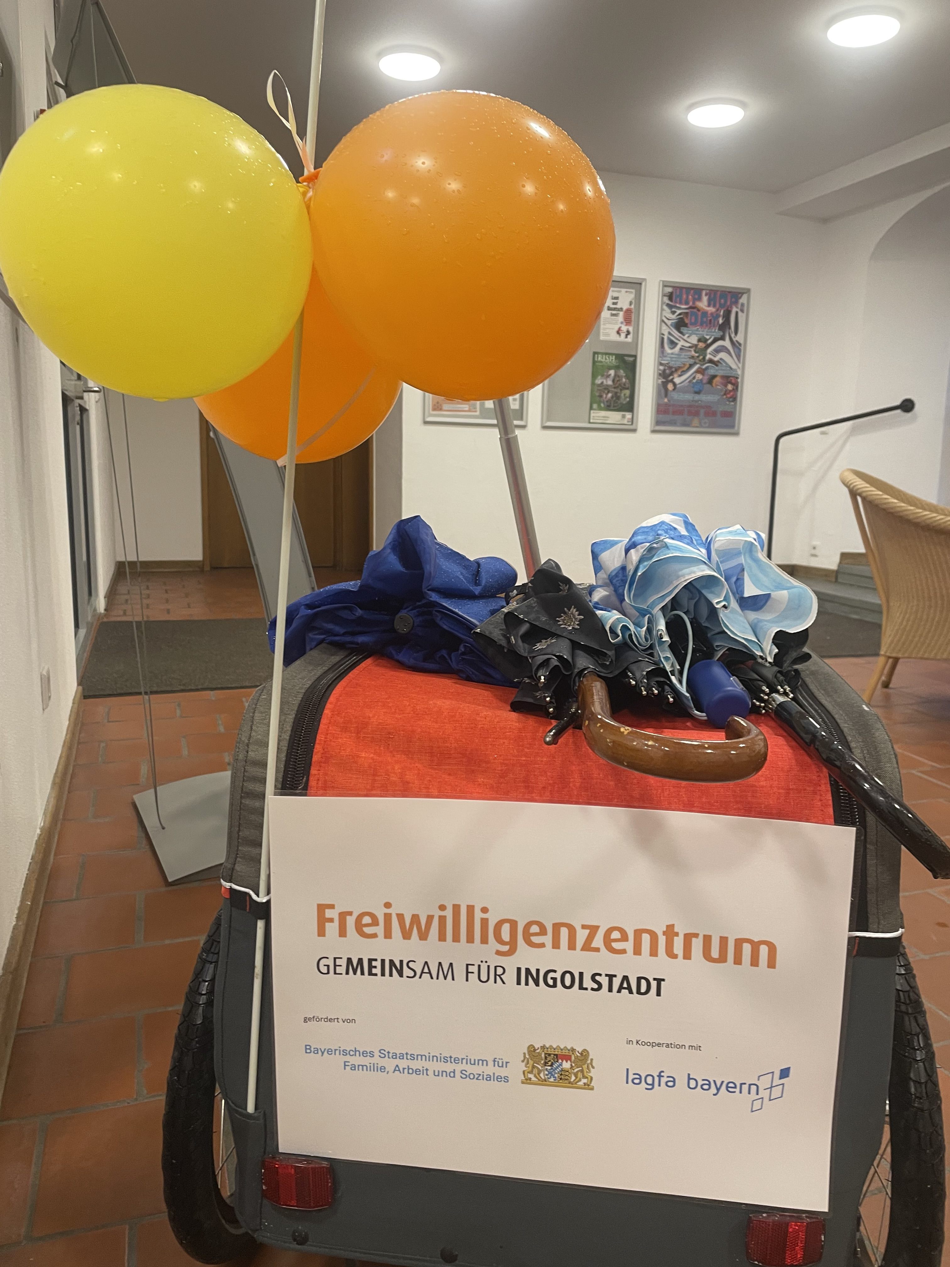 Auf dem Foto sieht man einen Bollerwagen von hinten, darauf liegen Schirmer, ein paar Luftballons sind am Wagen festgemacht. Am Wagen ist ein Plakat befestigt, mit dem Logo des Freiwilligenzentrums Ingolstadt sowie den Logos des Bayerischen Sozialministeriums sowie lagfa bayern e.V, als Förderer/Unterstützer